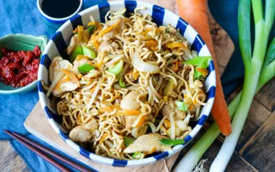 Bami Goreng mit Hühnchen, Karotten und Frühlingszwiebeln in einer Schüssel, daneben Sojasauce, Chili und frisches Gemüse.