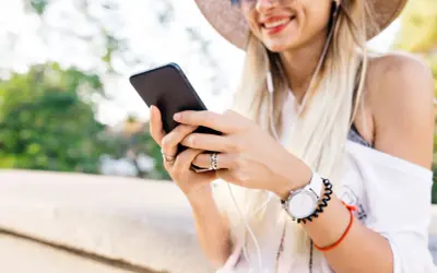 Uśmiechnięta kobieta w kapeluszu słucha muzyki przez słuchawki, trzymając telefon. Może szuka instrukcji, jak zainstalować radio w telefonie.