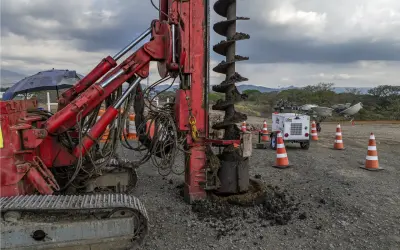 Máquina perforadora roja en obra, extrayendo tierra y formando columnas de grava. Conos naranjas y equipo de iluminación al fondo.