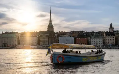A blue sightseeing boat with a yellow striped canopy sails on water in Stockholm, with historic buildings and a church spire in the background at sunset.