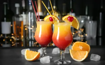 Dwa drinki Tequila Sunrise, ozdobione pomarańczą i wiśnią, stoją na barze. W tle widać butelki alkoholu i migoczące światełka. Idealne na wieczór z przyjaciółmi, jak w najlepszych lokalach Baczewski.