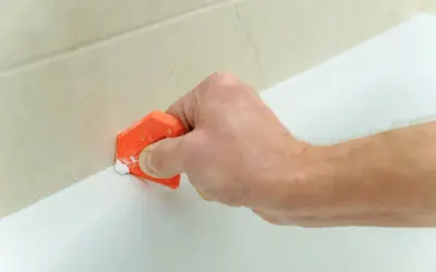 Mano usa spatola arancione per rifinire silicone, un lavoro antisilicone fai da te per sigillare la vasca.