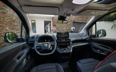 Interior de la nueva Citroën Berlingo, con pantalla táctil, volante moderno y asientos cómodos. Afuera, una pareja se acerca a un punto de carga.