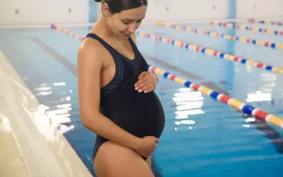 Kobieta w ciąży w czarnym stroju kąpielowym stoi przy basenie, delikatnie dotykając brzucha.