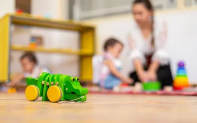 Drewniany krokodyl na podłodze żłobka. W tle opiekunka z dziećmi. Ciekawe, ile zarabia opiekun w żłobku państwowym za taką pracę.