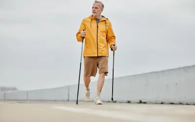 Mężczyzna w żółtej kurtce i brązowych spodenkach wykonuje rozgrzewkę przed nordic walking, trzymając kije.
