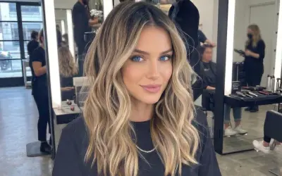 Kobieta z pięknymi, falowanymi blond włosami, z subtelnym, ciemnym odrostem przy blondzie. W tle salon fryzjerski.