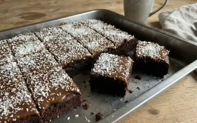 En kladdkaka med glasyr och kokosflingor skuren i bitar i en plåt. En mugg kaffe står bredvid.