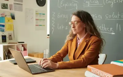 Nauczycielka w okularach pracuje przy laptopie. Na tablicy napisy po angielsku. To obraz awansu zawodowego nauczyciela, który rozwija swoje umiejętności.