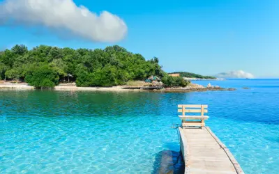 Albania - wczasy samolotem all inclusive. Drewniane molo z ławką prowadzi do krystalicznie czystej wody. Zielona wyspa w tle.