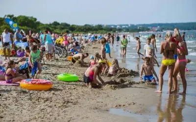 Tłoczna plaża jelitkowo w słoneczny dzień. Dzieci budują zamki z piasku, dorośli odpoczywają, a fale delikatnie obmywają brzeg.