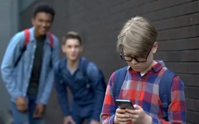 Chłopiec w okularach patrzy w telefon, podczas gdy dwaj inni chłopcy śmieją się za nim. Stop hejt!