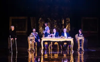 Scena z "Dziadów" w warszawskim teatrze. Przy złotym stole siedzą mężczyźni w strojach z epoki, obok kobieta na wózku.