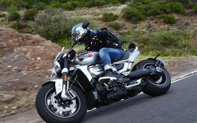 Motocyklista w kasku i kurtce jedzie na motocyklu Triumph Rocket 2 po krętej drodze wśród zieleni.