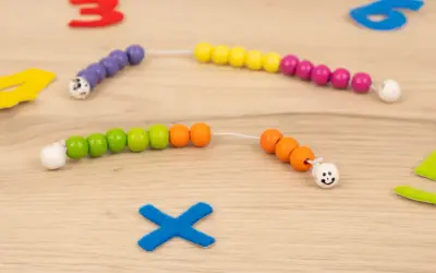 Bunte Perlenketten zum **rechenkette basteln** liegen auf Holz. Zahlen und ein blaues Kreuz sind im Hintergrund.