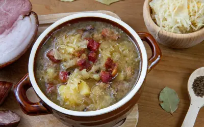 Gęsty, sycący kapuśniak z kawałkami boczku i ziemniaków, podany w glinianej misce. Obok miseczka z kiszoną kapustą i drewniana łyżka z kminkiem.