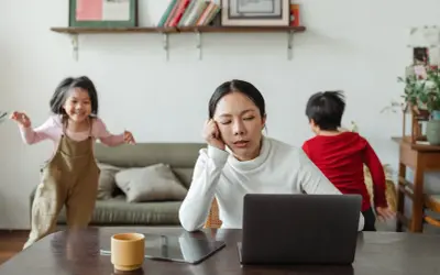 Donna esausta davanti al laptop, mentre i bambini giocano. Un quadro di **parental burnout** in casa.
