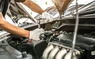 Mechanik w białych rękawiczkach dokręca śrubę kluczem nasadowym. Silnik samochodu jest otwarty, co sugeruje przegląd samochodu po terminie.
