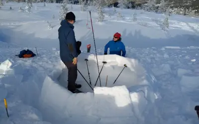 Två personer bygger en bivack i snön. De använder snöblock för att bygga väggarna och stavar för att markera utrymmet.