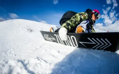 Snowboardzista w kamizelce moro i różowej czapce, zjazd na desce. Warto wiedzieć, jak dobrać buty snowboardowe, by czuć się pewnie na stoku.