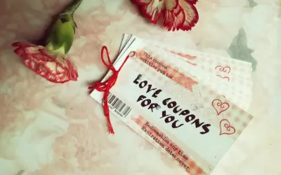 Papierowa rocznica uczczona kuponami miłości. Dwa kwiaty goździka i stosik kuponów "Love Coupons for You" na różowym tle.