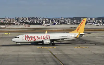 Samolot linii lotniczych Pegasus z napisem "flypgs.com" na kadłubie na płycie lotniska, w tle miasto.
