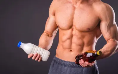 Muskulöser Mann hält Milch und eine Flasche mit bernsteinfarbener Flüssigkeit. Die Frage: Alkohol und Muskelaufbau – eine gute Kombination?