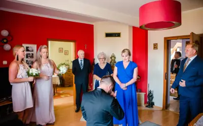 Mężczyzna klęka, całując dłoń kobiety. Rodzina zgromadzona w pokoju, oczekując na prośbę o błogosławieństwo.