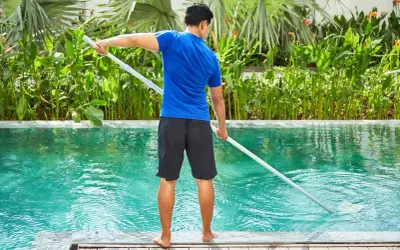 Hombre con camiseta azul y pantalones cortos negros limpia piscina de sal con un limpiafondos.