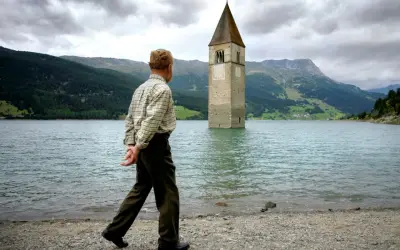 Ein Mann spaziert am Ufer eines Sees, auf dem ein Kirchturm aus dem Wasser ragt. Die Kirche im Stausee ist ein eindrucksvolles Zeugnis der Vergangenheit.