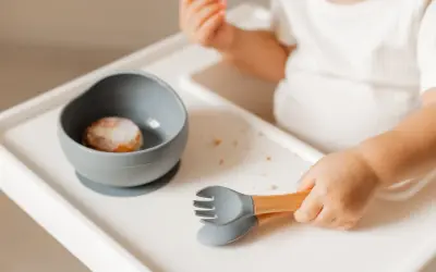 Małe rączki chwytają sztućce, gdy dziecko samo je. Miseczka z jedzeniem i okruszki na tacce.