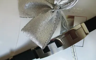 Elegancka bransoleta z czarnym paskiem i metalowymi elementami, ozdobiona srebrną kokardą. Idealnie pokazuje, jak zapakować biżuterię na prezent.