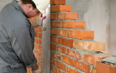 Murarz w białym kasku sprawdza pion muru, używając poziomicy. Widać cegły o standardowych wymiarach, układane na zaprawie.