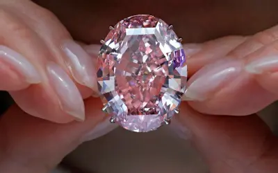Dłonie trzymają olśniewający, owalny, różowy diament, prawdopodobnie najdroższy diament na świecie, błyszczący w świetle.