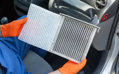 Ręce w pomarańczowych rękawiczkach pokazują brudny filtr kabinowy Toyoty Yaris, gotowy do wymiany.