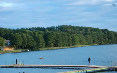Spokojny dzień nad jeziorem Sierakowo. Ludzie spacerują po pomoście, a na wodzie pływają kajaki.