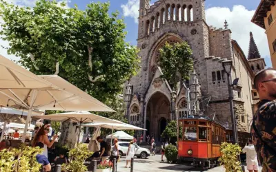 En charmig spårvagn passerar framför en kyrka i Soller, Palma. Folk sitter vid uteserveringar under parasoll.