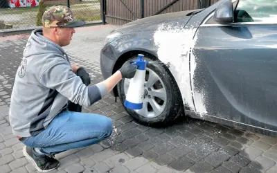 Mężczyzna w czapce z daszkiem i jeansach kuca przy samochodzie, rozpylając pianę na błotnik. Czy można naprawiać auto pod blokiem? Tak, jeśli to drobna naprawa lakieru.