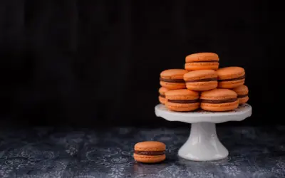 Pomarańczowe makaroniki francuskie z czekoladowym nadzieniem ułożone na białej paterze, jeden makaronik leży obok.