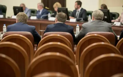 Mężczyźni w garniturach siedzą przy stole, czekając na **absolutorium**. W tle widać monitory i innych uczestników spotkania.