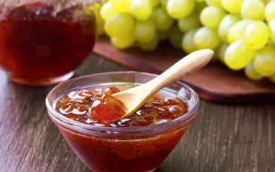 Un cuenco de cristal con mermelada de uvas casera, una cuchara de madera y un racimo de uvas verdes al fondo.