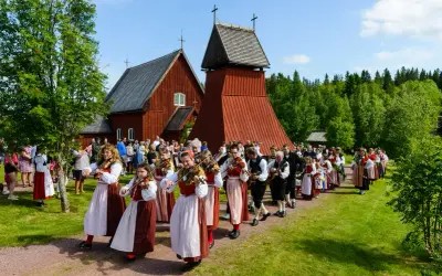 En procession av musiker i folkdräkter, som spelar fiol och gitarr, går förbi en röd träkyrka. Många människor i traditionella kläder, vilka firar midsommar, följer efter.