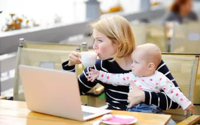 Kobieta pije kawę, trzymając dziecko. Obok leży laptop. Karmienie piersią i chwila dla siebie.