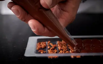 Main d'un artisan versant du chocolat fondu dans un moule pour créer une tablette de chocolat maison garnie de noisettes caramélisées.
