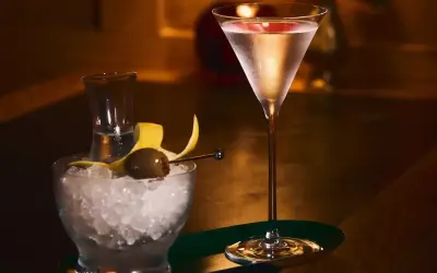 Elegancki drink z martini w kieliszku, obok miseczka z lodem, oliwką i skórką cytryny.