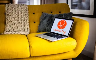 Żółta kanapa z laptopem otwartym na stronie couchsurfing. Obok koc i szara poduszka.