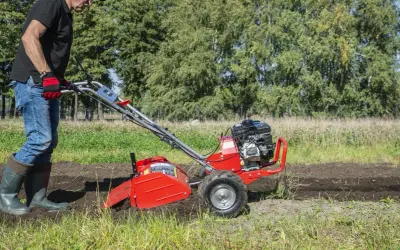 Mężczyzna w rękawiczkach i gumowcach obsługuje czerwoną glebogryzarkę na polu.