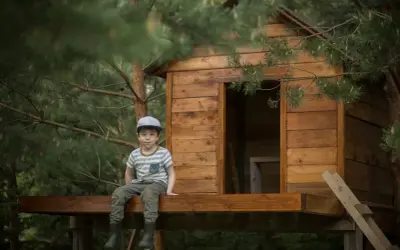 Chłopiec w czapce siedzi na tarasie domku na drzewie. To inspiracja, jak zrobić domek na drzewie dla dzieci.