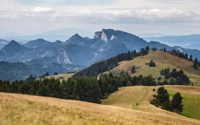 Malowniczy krajobraz górski, idealny na trasę Szczawnica. W oddali widać skaliste szczyty, a na pierwszym planie zielone wzgórza i ścieżkę dla turystów.