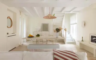 Un soggiorno luminoso con soffitto a travi e **bianco caldo pareti**. Tavolo da pranzo in legno, sedie, panca e una scala bianca conducono a un piano superiore.